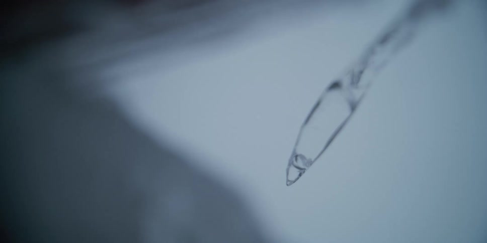 An extreme close-up slow-motion shot shows a melting icicle with a water droplet forming at the tip