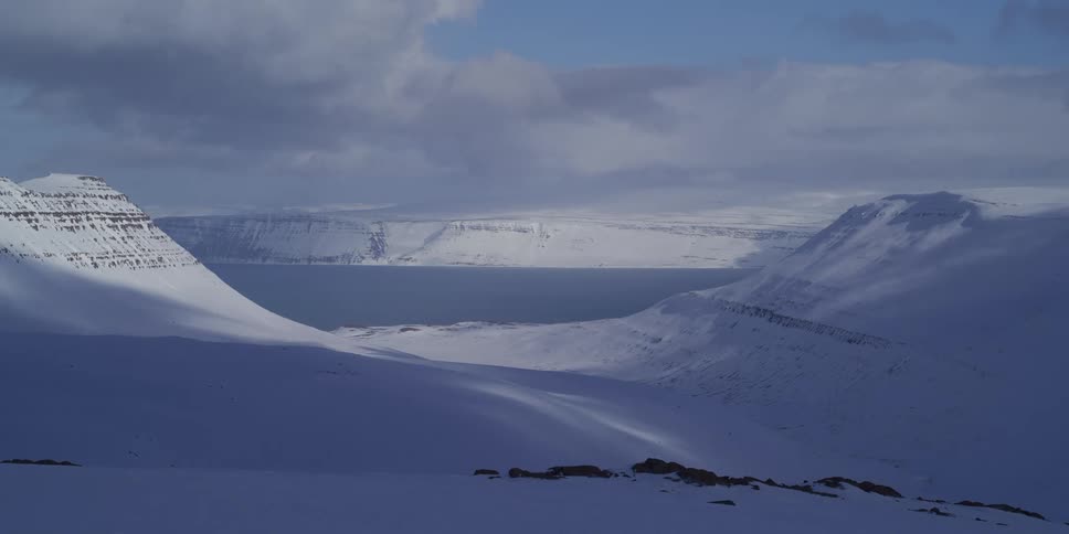 A breathtaking wide shot reveals the vast, snow-covered mountains and valley of Iceland's Westfjords leading out to the serene ocean