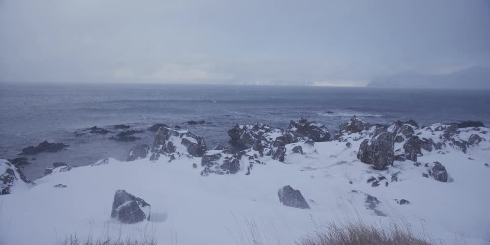 Strong winds blow snow across a frozen, rocky shoreline during a winter storm