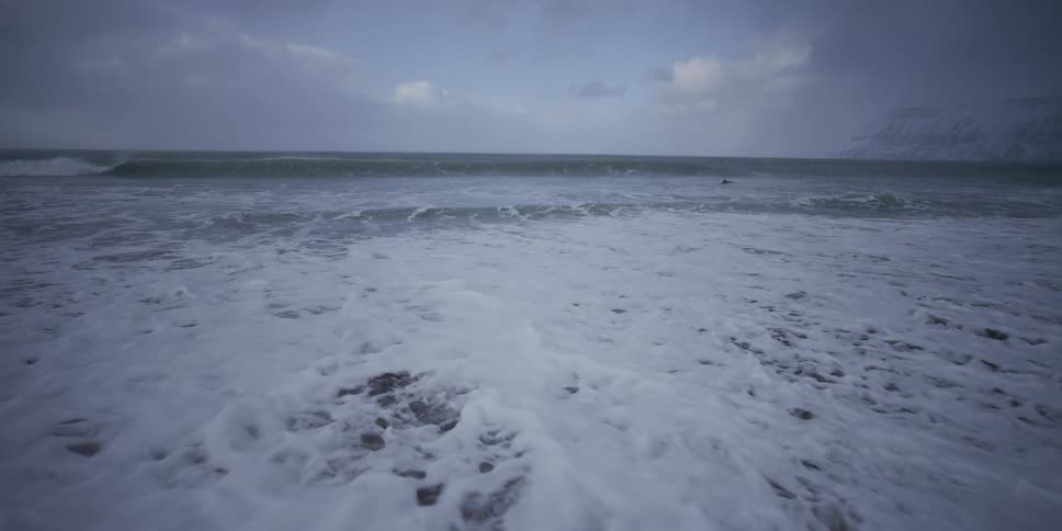 Foamy waves recede from a dark, snowy shoreline in super slow motion on a cold winter day