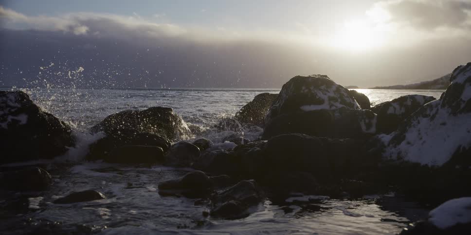 Winter ocean waves crash in slow motion against snow-dusted rocks as the sun sets