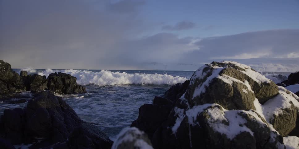 A powerful ocean wave rolls towards a snow-covered rocky coastline in a majestic slow-motion shot