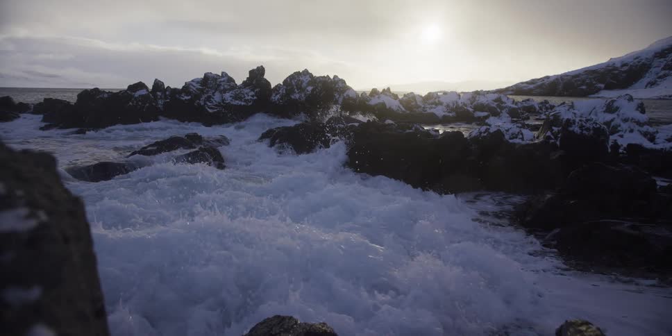 Powerful ocean waves crash over a snow-covered rocky coastline in dramatic slow motion