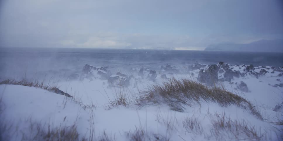 A harsh winter blizzard blows snow across a desolate, rocky coastline in a dramatic slow-motion shot