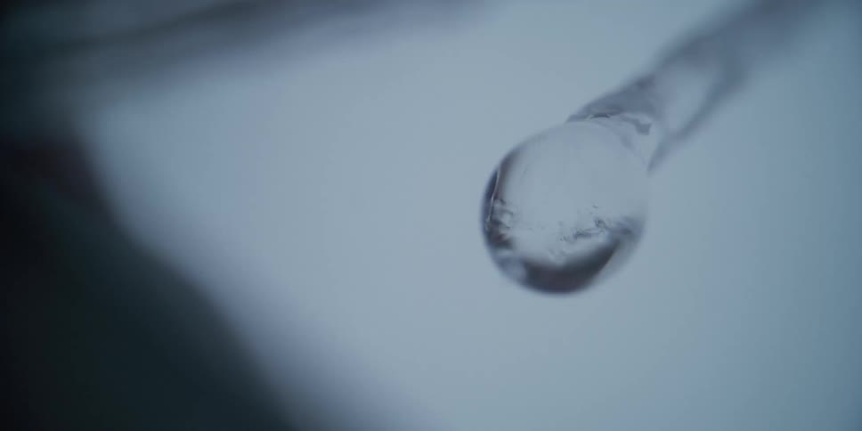 A macro close-up shot shows a single drop of water forming on the tip of a melting icicle before dripping away