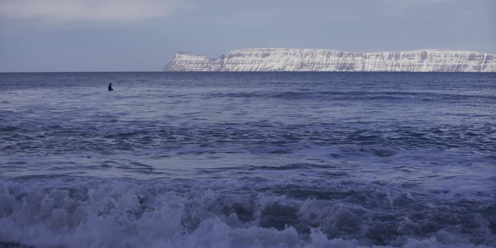 Waves crash in slow motion as a surfer waits in the icy ocean with a vast snow-covered cliff in the distance