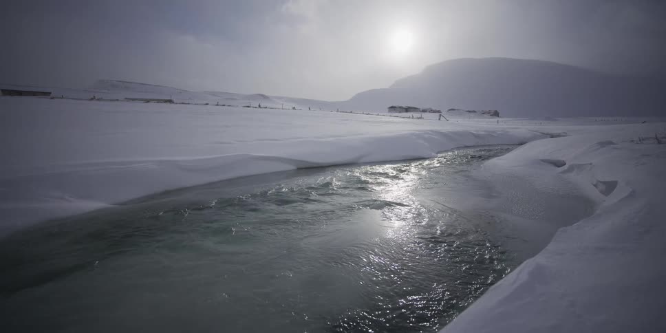 Sunlight shimmers on an unfrozen river flowing through a vast, snowy landscape in slow motion
