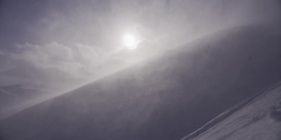 A powerful winter blizzard whips snow across a steep mountain slope as the sun tries to break through the clouds