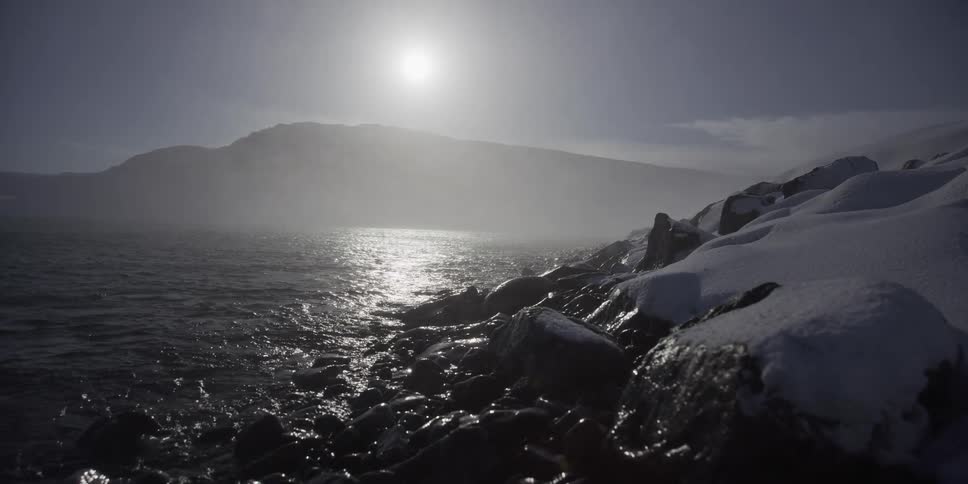 A slow slider shot reveals a stark, snowy shoreline with misty water reflecting the low winter sun