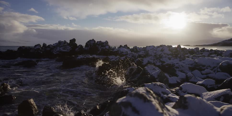 Slow motion waves crash against snow-covered rocks on a cold, sunny coastline during winter