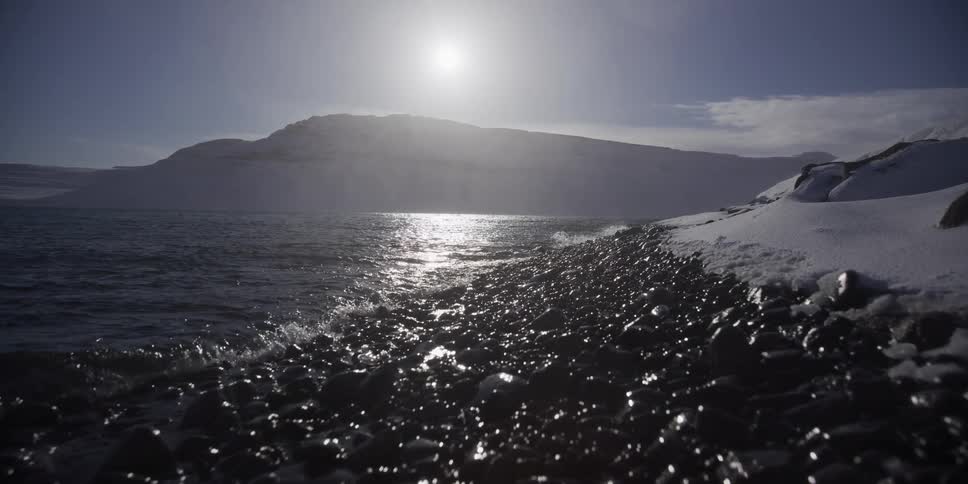 Slow-motion waves gently wash over a rocky, snow-covered shore under a bright, cold sun in the Arctic