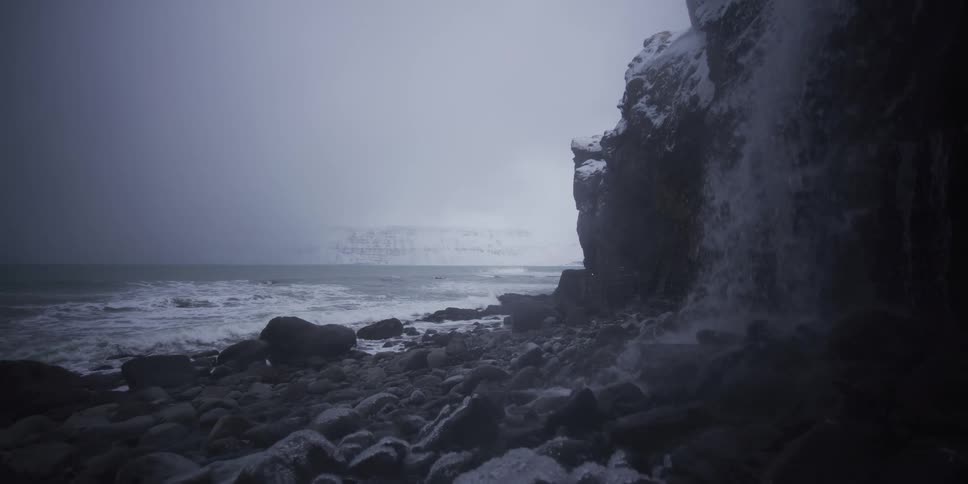 A slow-motion view from behind a waterfall shows a desolate snowy coastline as ocean waves crash onto the rocky shore