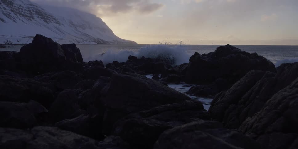 An ocean wave breaks in dramatic slow motion against a rugged, rocky shoreline backed by snow-covered mountains at dusk