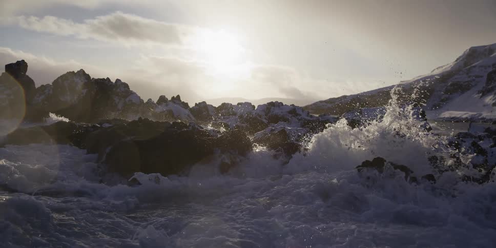 Powerful ocean waves crash in slow motion against a snow-covered rocky coastline as the sun sits low in the sky