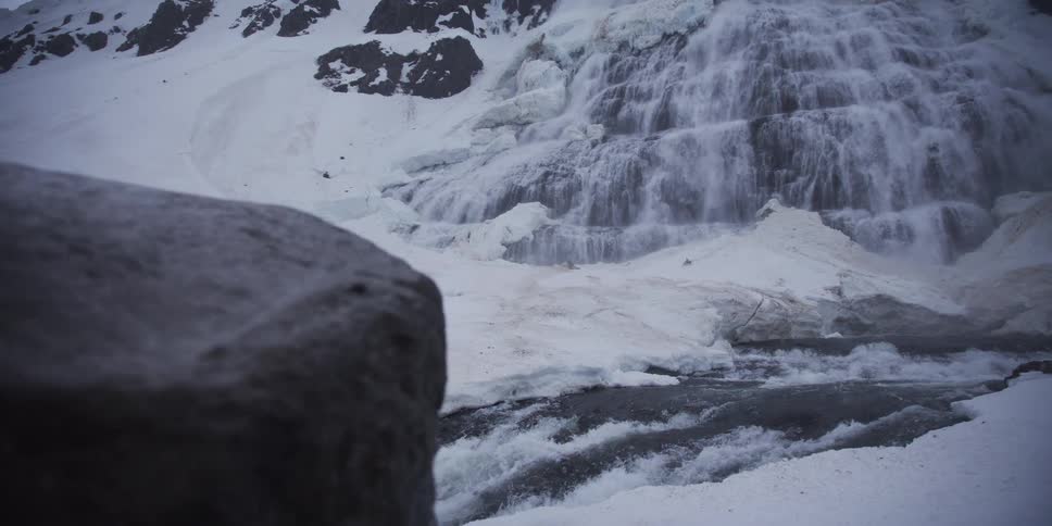 A powerful, icy waterfall cascades down a snow-covered mountain into a rushing river, revealed in a slow pan from behind a rock