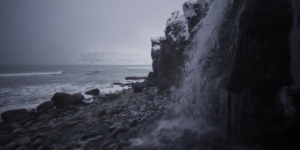 A waterfall cascades in slow motion from a snow-covered cliff onto a rocky shore beside the cold ocean