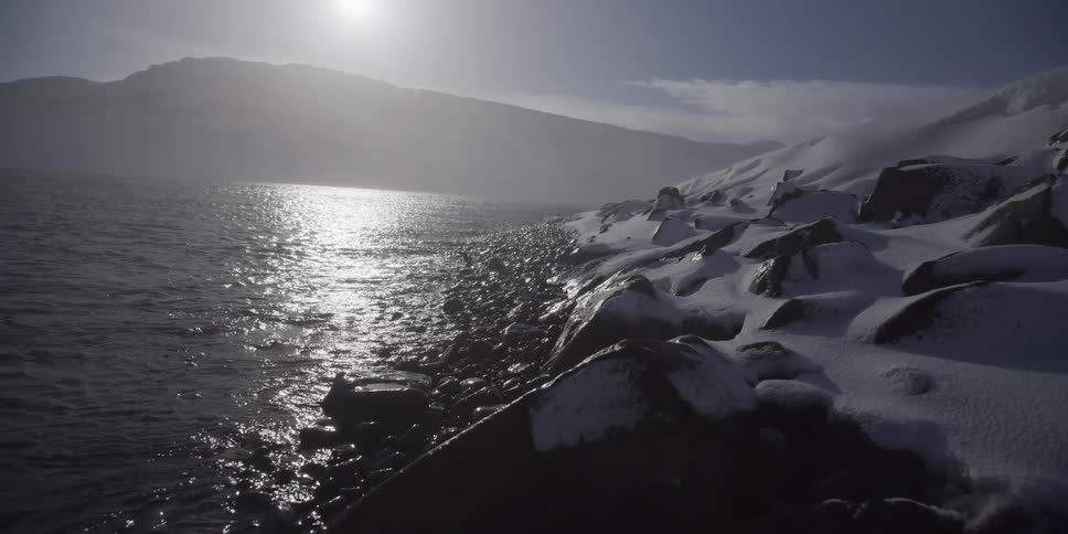 A slow pull back reveals a stark, snow-covered rocky coastline as water shimmers under the bright sun in a frigid, desolate landscape