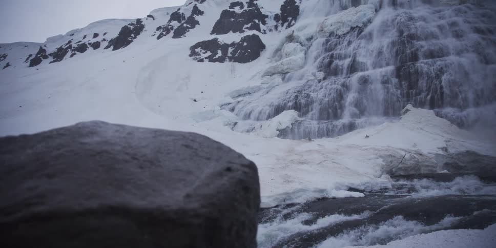 A slow dolly shot reveals a powerful, partially frozen waterfall cascading down a snowy mountain landscape