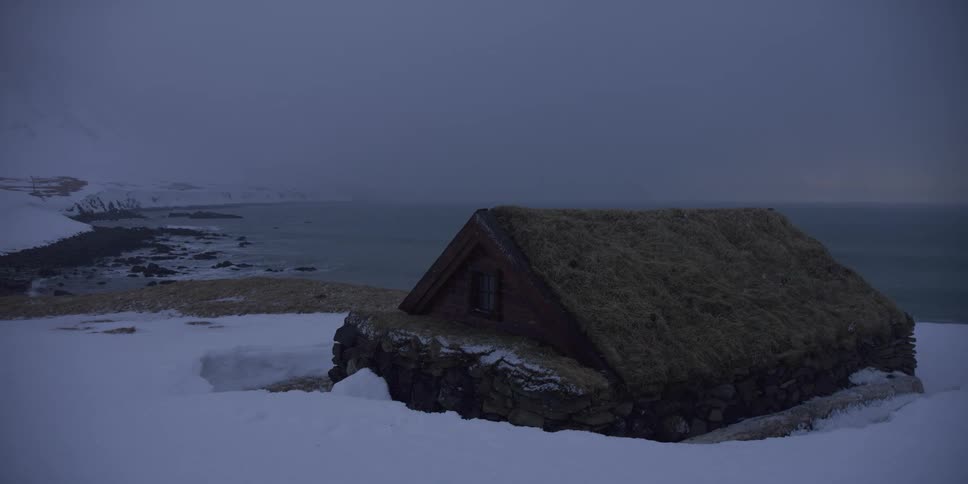 A traditional Icelandic turf house sits alone on a snowy cliff overlooking the cold ocean on an overcast day
