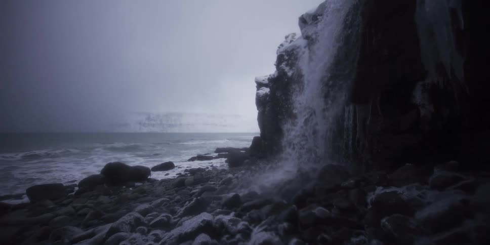 Slow motion captures a waterfall cascading onto a desolate, snowy beach beside a frigid winter sea