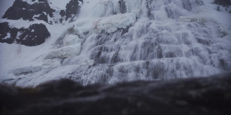 A powerful waterfall flows through snow and ice on a rugged mountainside in winter, viewed from a low angle