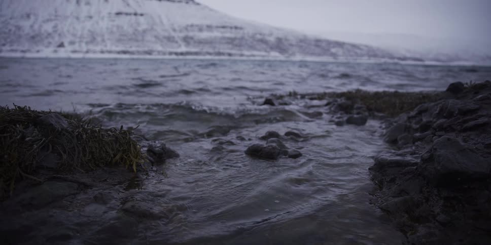 Slow motion close-up of gentle waves washing over a rocky shoreline in a snowy Icelandic fjord during winter