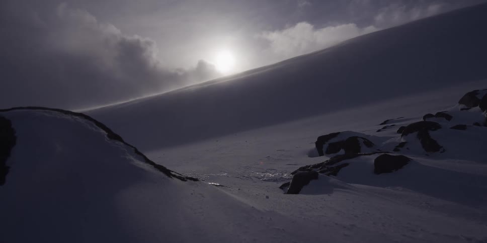 A bleak, windy, and snow-covered mountain landscape is backlit by the sun shining through a cloudy sky