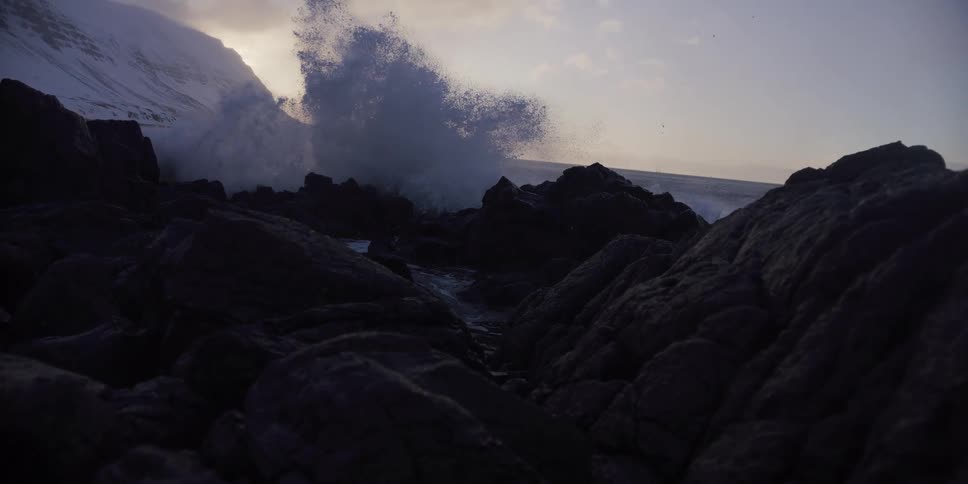 In dramatic slow motion, a powerful ocean wave explodes against dark volcanic rocks on a winter coastline