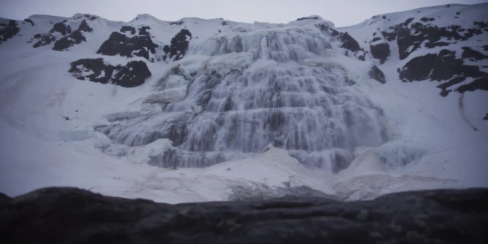 A majestic tiered waterfall thunders down a snow-covered mountain, surrounded by ice and stark rocky terrain