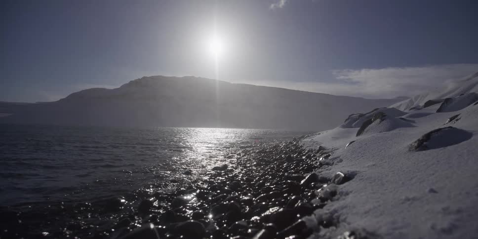 A slow dolly shot pulls back along a snowy, rocky shoreline as the bright arctic sun reflects on the calm water