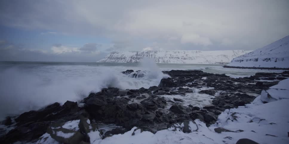 A powerful ocean wave crashes in slow motion against a rocky, snow-covered shoreline with majestic mountains in the distance