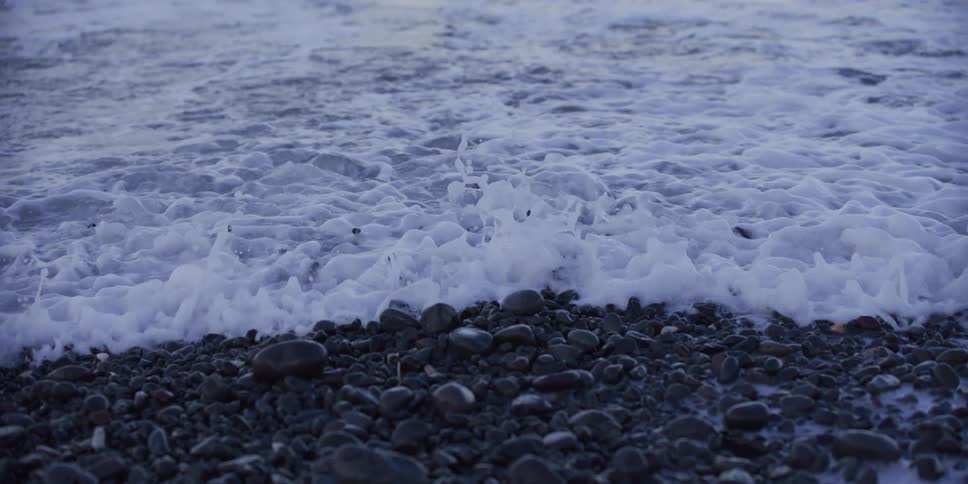 A slow-motion close-up captures the gentle foam of ocean waves washing over a dark, pebbled shoreline