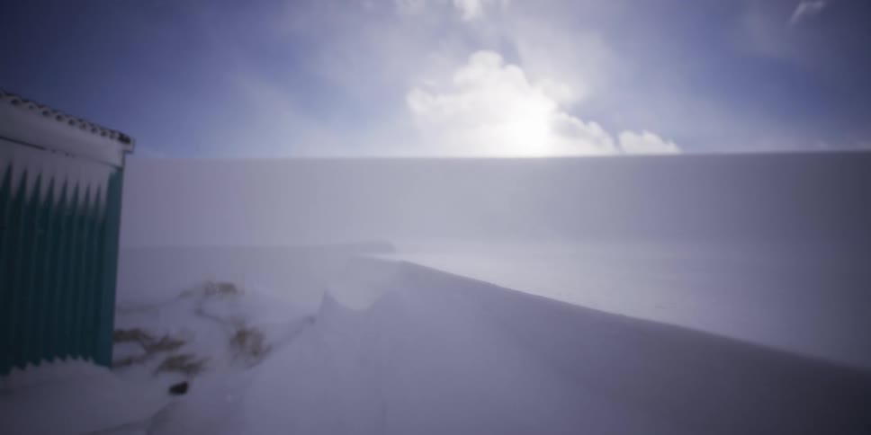 Heavy winds create a blizzard over a snow-covered landscape, revealing a large snowdrift next to a remote shed as the camera pulls back