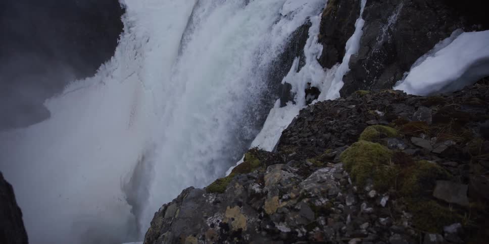 A powerful waterfall crashes down a snowy, rocky cliff in a dramatic slow-motion close-up shot