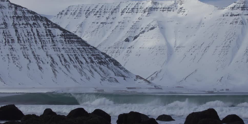 A majestic ocean wave breaks in slow motion with towering snow-covered mountains in the background