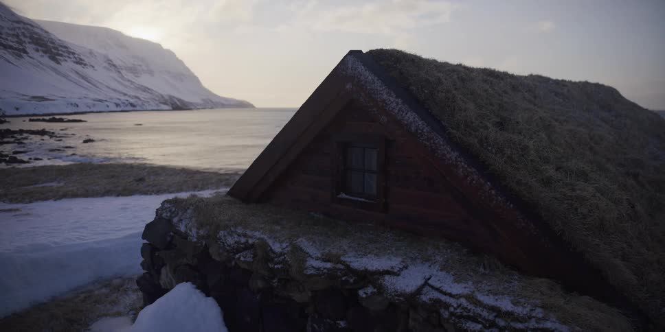 A traditional turf-roofed house stands on a snowy shoreline in Iceland, with the calm ocean and mountains in the background