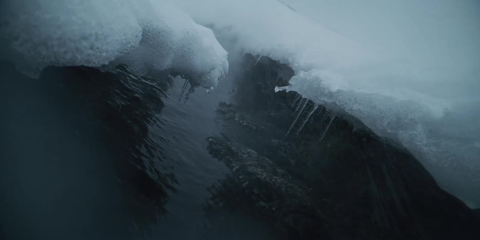 Eerie steam rises from a dark, rippling river as icicles melt along the snow-covered bank