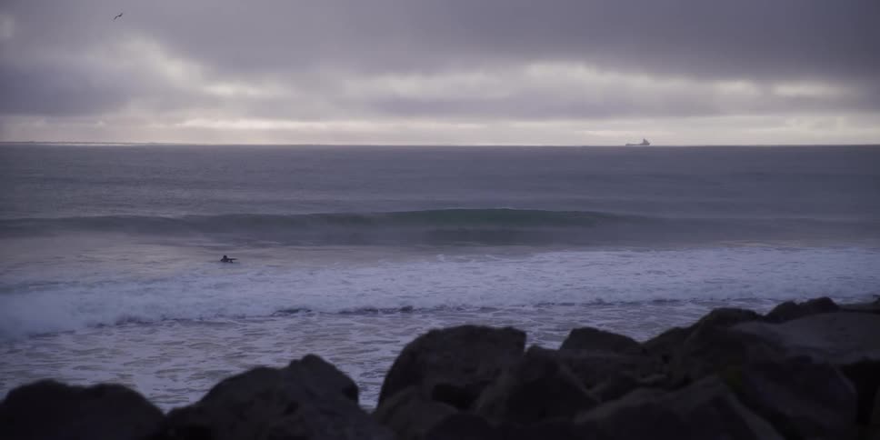 From behind coastal rocks, a solitary surfer waits as a perfect wave forms and breaks in slow motion on a moody, overcast morning