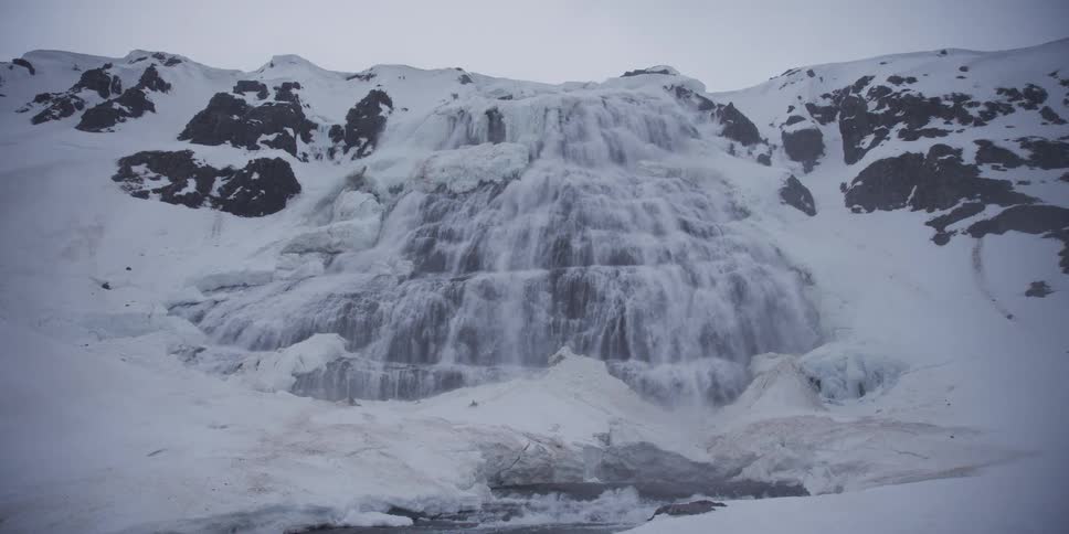 A slow, rising dolly shot reveals a majestic, partially frozen waterfall cascading down a vast, snow-covered mountain