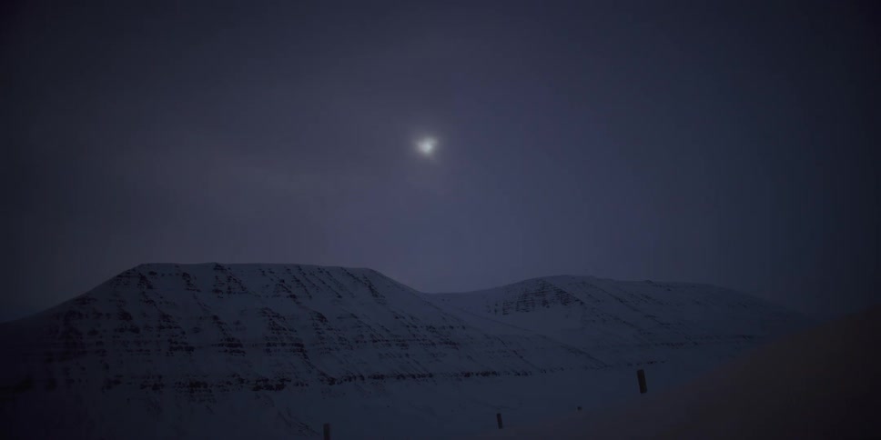 Ethereal time-lapse of a bright moon moving through the night sky and behind clouds, casting light over a serene, snow-covered mountain landscape