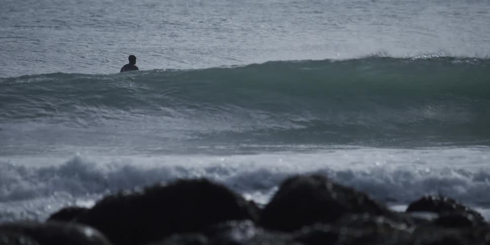 A silhouetted surfer paddles over a rising ocean swell to catch a breaking wave in dramatic slow motion