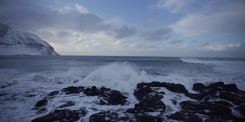A powerful ocean wave crashes in dramatic slow motion against a snow-covered rocky coast