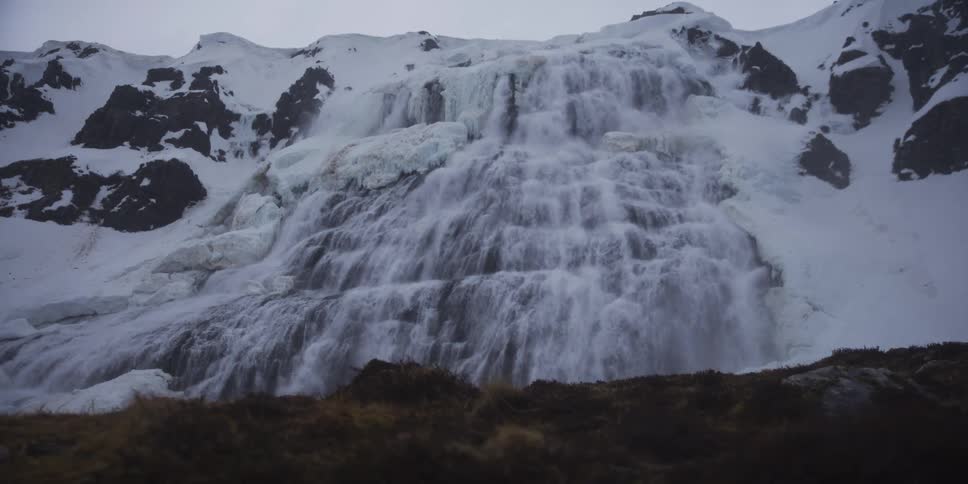 A powerful waterfall rushes down the side of a snowy, icy mountain on an overcast day