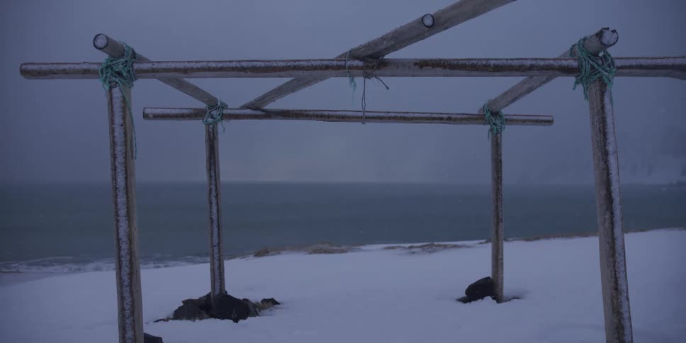 Snow gently falls on a frozen, desolate arctic beach with empty fish drying racks overlooking the cold ocean