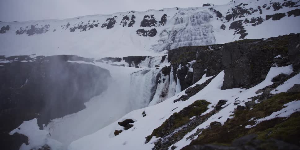 A stunning, wide dolly shot showcases a powerful waterfall cascading through a vast, snow-covered Icelandic canyon in winter