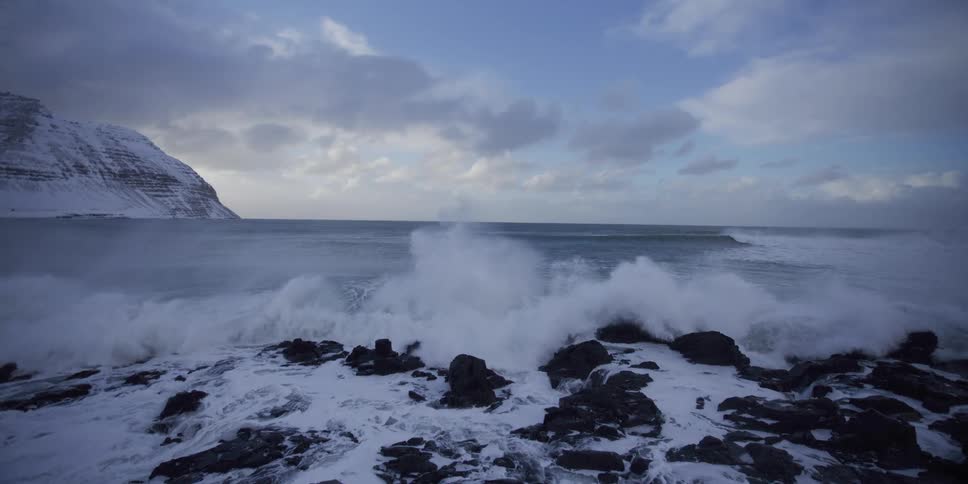 Dramatic slow motion shot of powerful ocean waves crashing against a snowy, rocky shoreline in a winter landscape