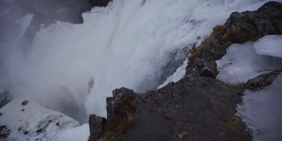 A dramatic slow-motion push-in shot over a snowy, rocky cliff edge reveals a powerful, churning waterfall