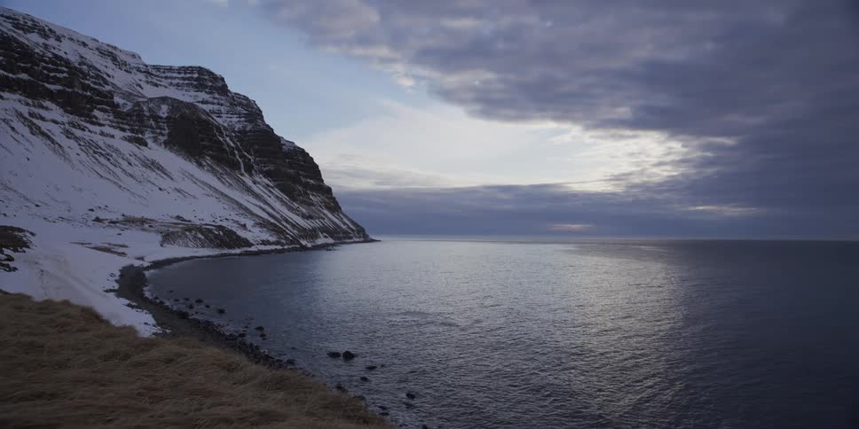A majestic snow-covered mountain meets the calm ocean along a rocky coastline under a dramatic, cloudy sky