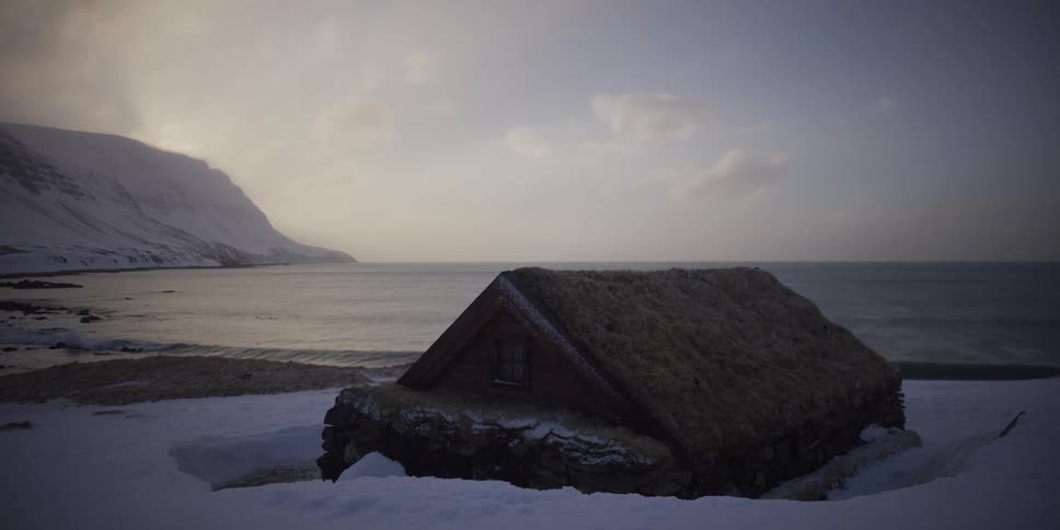 A quaint turf-roof cabin sits on a snowy beach next to a tranquil fjord in a serene winter landscape