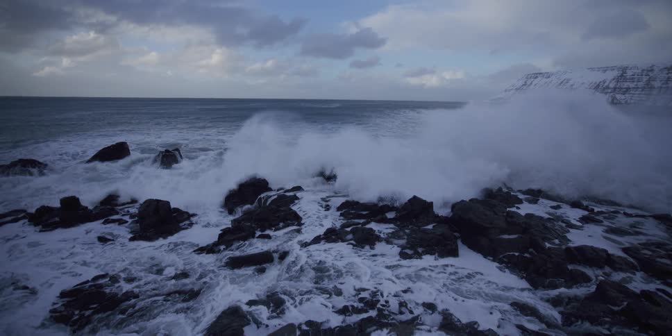 The raw power of the ocean is on full display as a massive wave crashes in slow motion over a snowy, rocky shoreline